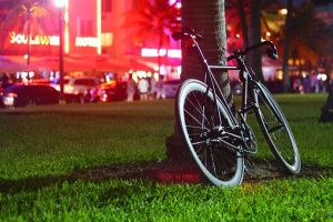 Bicycle leaned on a palm tree, overlooking a busy street at night. Choose between best neighborhoods for expats in North Miami Beach