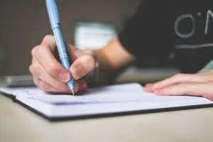 A close up of a person holding a blue pen and writing something in a notebook. You need to know exactly what not to put in your storage unit, so you should pay attention to the list of items in front of you.