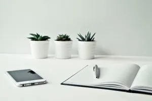 An open notebook, a white phone, and white flower pots on a white desk.