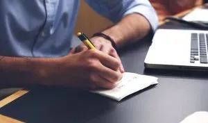 A close up of a man in a blue shirt holding a pen and writing something on a piece of paper. He might be making an office relocation checklist, which will help him move his business to a different location.